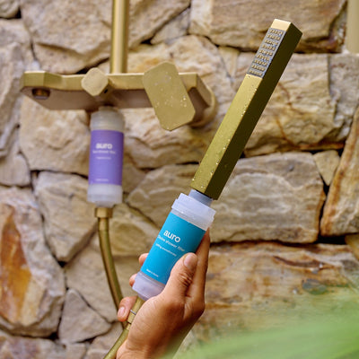 Person holding a shower head with a Suuqo water filter against a stone wall background
